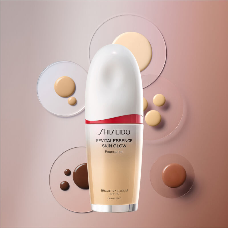 A bottle of the Shiseido Revitalessence Skin Glow Foundation SPF 30 against a brown background, dotted with several small circles of foundation in different colors to show its serum-like texture that is available in a wide range of shades.