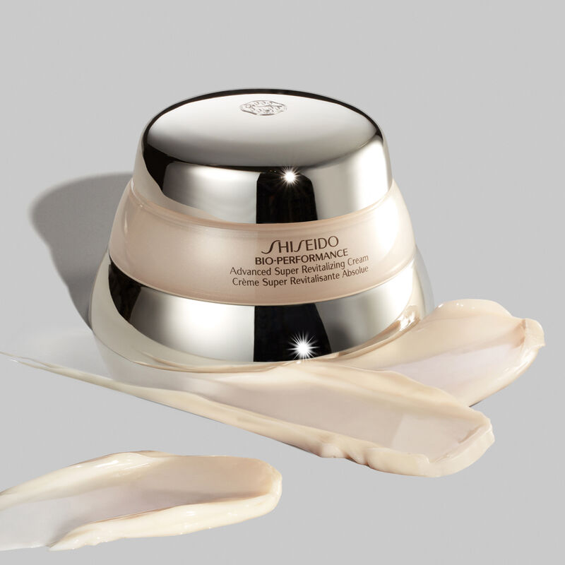 "Un frasco de la crema Shiseido Bio-Performance Advanced Super Revitalizing Cream sobre un fondo gris. Hay varias muestras de la crema con textura rica delante del frasco".