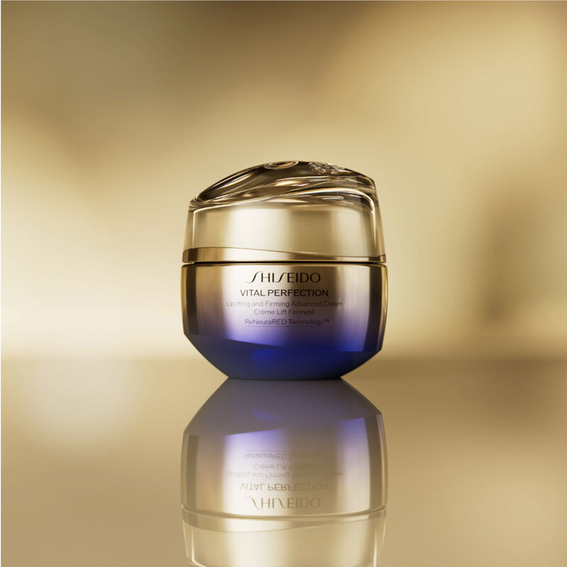 Un frasco redondo, cerrado, de color dorado y morado, de la crema Shiseido Vital Perfection Uplifting and Firming Advanced Cream. El frasco se muestra sobre un fondo dorado difuminado.