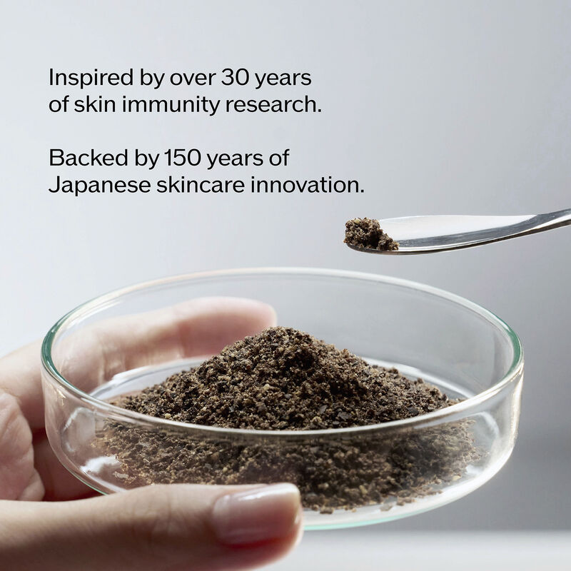 Texto: "Inspirado en más de 30 años de investigación sobre inmunidad de la piel. Respaldado por 150 años de innovación en tratamiento japonés". En la parte inferior, una mano sostiene una placa de Petri con un polvo oscuro y granular; encima, una cuchara con una pizca del polvo.