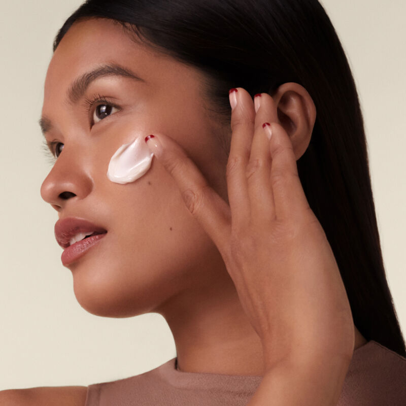 "Una modelo aplicándose la crema Shiseido Bio-Performance Advanced Super Revitalizing Cream en la mejilla mientras mira hacia otro lado, mostrando la rica textura de la crema".