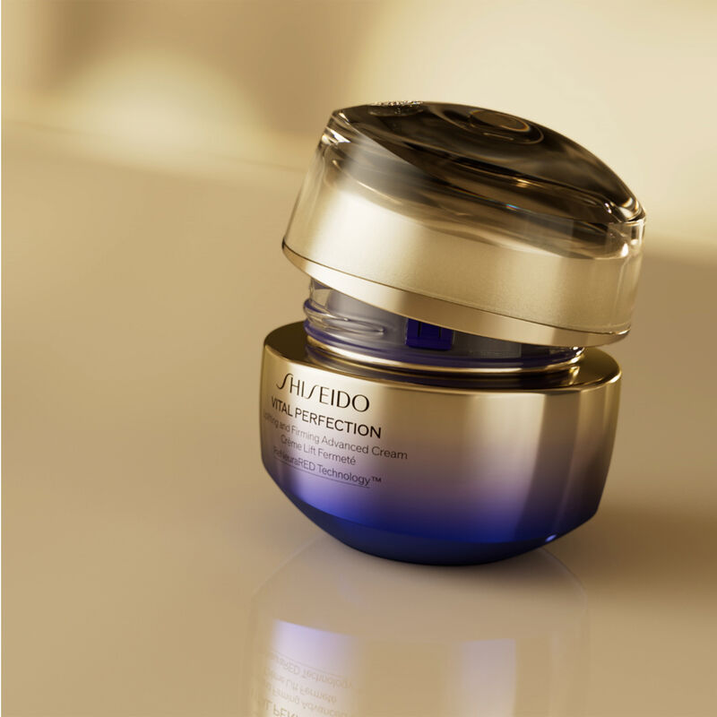 Un frasco redondo, medio abierto, de color dorado y morado, de la crema Shiseido Vital Perfection Uplifting and Firming Advanced Cream. El frasco se muestra sobre un fondo dorado difuminado.