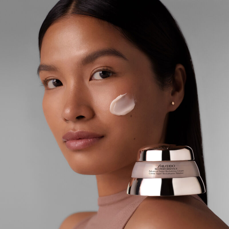 "Una modelo con la crema Shiseido Bio-Performance Advanced Super Revitalizing Cream aplicada en la mejilla mientras mira hacia la cámara, mostrando la rica textura de la crema. El frasco del producto también está sobre su hombro izquierdo".